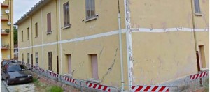 Exterior view of a damaged building with visible cracks, surrounded by construction barriers, indicating it is uninhabitable.